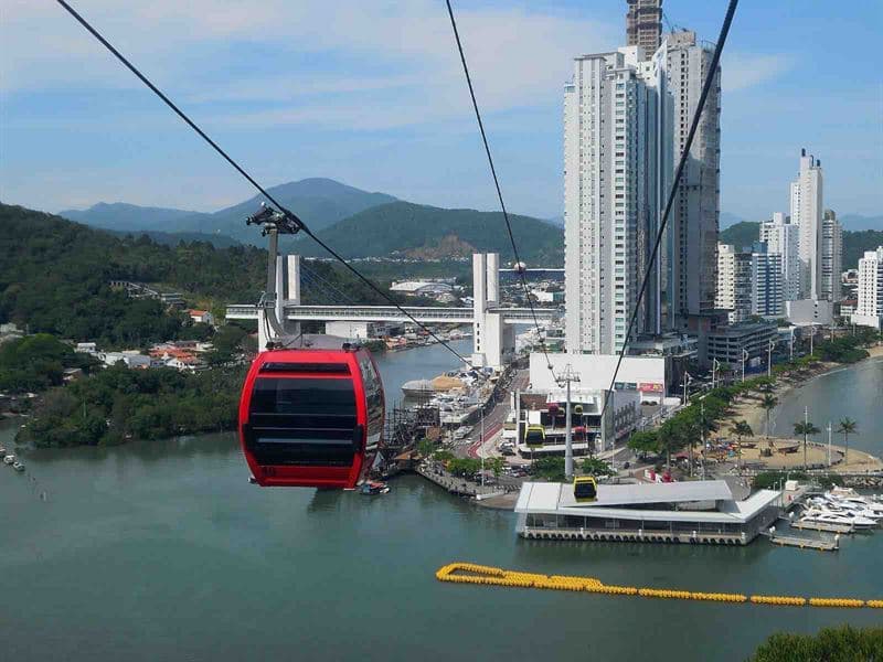 Bondinho vermelho do Parque Unipraias sobre o rio, com os prédios altos de Balneário Camboriú ao fundo.